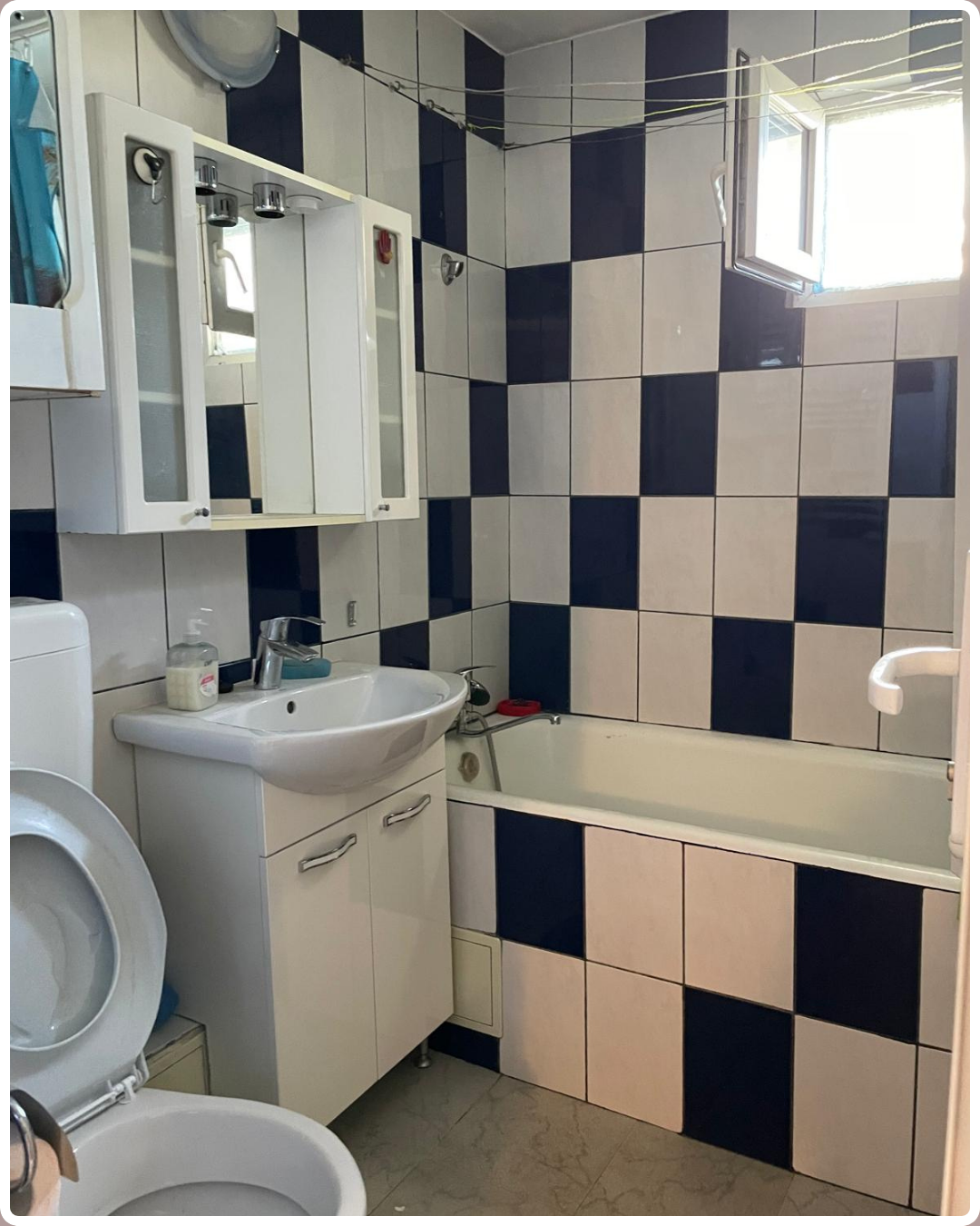 Baie veche cu faianță alb-bleumarin în damă, mobilier deteriorat și sârmă pentru rufe — înainte de renovare